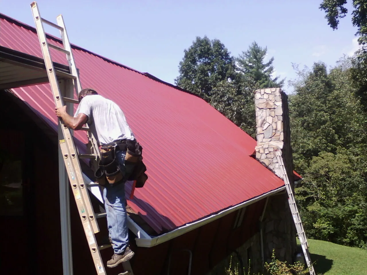 Roofer on a ladder beside a red metal roof and stone chimney — Soffit & Fascia Repair by Roofing Brothers in Watertown